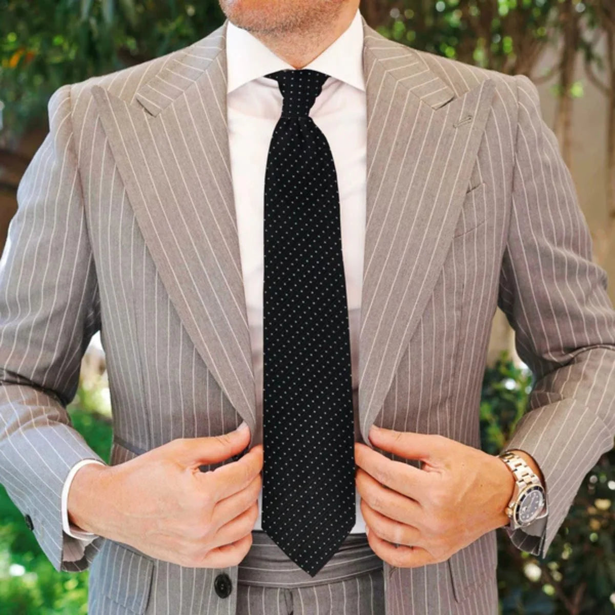 Man wearing a gray pinstripe suit with a black tie and watch, outdoors.